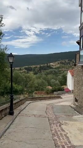 A vertical video shows a view down a cobblestone street lined with stone buildings on the right and a black lamppost on the left. In the background, a lush green valley is visible, with trees and rolling hills leading up to a distant mountain range under a cloudy sky. The camera is stationary.