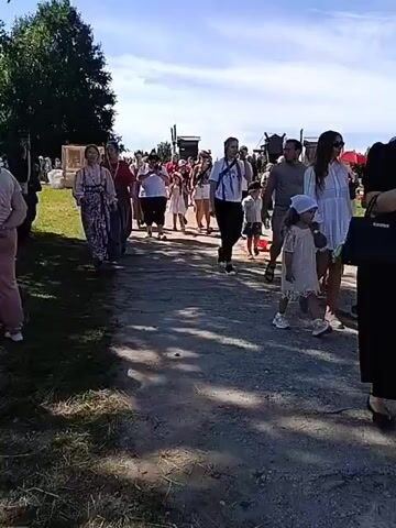 A group of people are walking down a dirt path on a sunny day. Some of the people are wearing traditional clothing. A few people are holding up phones, possibly recording or taking pictures. The path is lined with grass and trees. There are some structures in the background, possibly tents or booths, suggesting an outdoor event or festival.