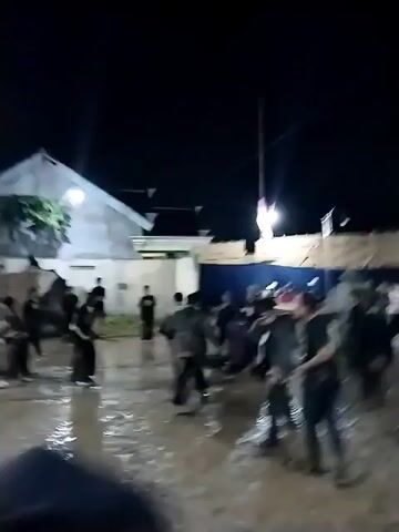 The video captures a night-time cultural performance or martial arts demonstration in Jenggrik, Indonesia. The scene is an open, unpaved area, with the ground heavily muddied and filled with puddles that reflect the artificial lighting. In the background, white-walled buildings with dark roofs are visible, along with some temporary wooden structures.

Numerous young men are present, mostly dressed in dark clothing. The central figures are actively engaged in dynamic movements, resembling a traditional Indonesian martial art like Pencak Silat or a vigorous dance. They wear black tops and pants, with several individuals adorned in colorful sarong-like fabrics featuring red, yellow, and white patterns tied around their waists or hips. One participant wears a red head covering. The performers exhibit energetic kicks, punches, and fluid body movements, appearing focused on their routine. Other individuals stand around the perimeter, observing the performance.

The area is illuminated by street lamps and other light sources, casting reflections on the wet ground. The activity is accompanied by music, suggesting a lively community event. The muddy conditions indicate either recent rain or a location commonly used for such performances. No visible text is discernible in the footage.