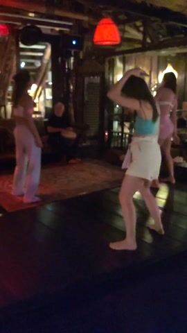 The video depicts a dimly lit interior space with a wooden floor and ceiling. A red light fixture hangs prominently from the ceiling. In the background, there's a man seated on a cushion, seemingly playing a drum. In the foreground, there are three women. One woman is wearing white pants and a light-colored top and is standing still. Another woman, dressed in white shorts and a turquoise top, is barefoot and actively dancing. A third woman, further back, is wearing a light pink outfit. There's a rug on the floor in the background. The room has a casual, bohemian vibe.