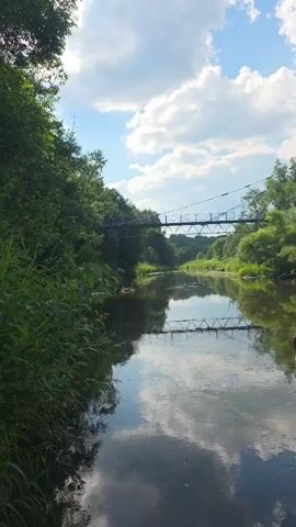 A camera pans across a calm river on a cloudy day. On the left, lush green foliage borders the water. In the distance, a suspension bridge spans the river, flanked by trees. The bridge and the surrounding greenery are reflected in the still water, which also reflects the white clouds in the blue sky. In the foreground, a few weathered wooden posts emerge from the water.