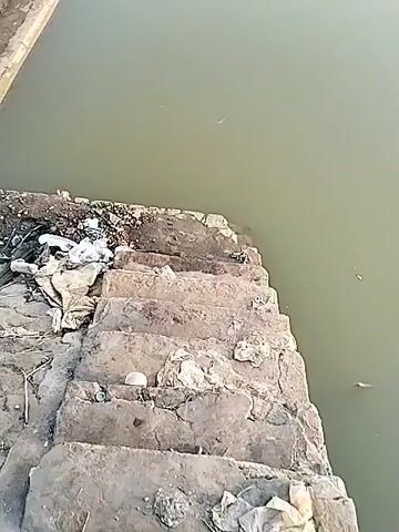 A first-person perspective video shows a concrete structure with steps leading down to a murky body of water. Trash is visible on the steps and the surrounding area. The camera then pans to show a wider view of the body of water, revealing a few people fishing from a concrete embankment in the distance. The sky is overcast, and the overall environment appears somewhat desolate.