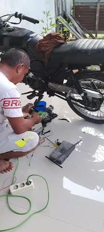 A man is kneeling on the floor, working on a black motorcycle. He is wearing a white t-shirt with red lettering and grey shorts. The motorcycle is partially disassembled, with parts laid out on the floor around it. There is a blue container and various tools visible. An electrical extension cord with multiple outlets is plugged into the wall, with wires connected to the motorcycle engine. The scene appears to be outdoors, possibly a driveway or open-air workshop, with a tiled floor and some vegetation in the background. The man's hands are actively engaged in manipulating components of the motorcycle.