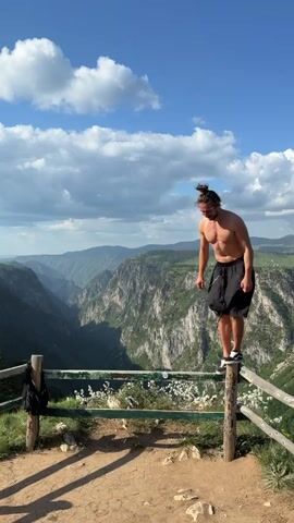 A man is seen standing on top of a weathered wooden fence overlooking a large canyon in Montenegro. He is fair-skinned and has shoulder-length brown hair pulled into a bun. He is shirtless but wearing black shorts and white shoes. He falls backward off of the fence and lands on his knees, unharmed. He then gives a thumbs up to the camera before it pans out to show the vast canyon and surrounding mountains. The sky is blue with fluffy white clouds. A small black bag is seen hanging on the fence to the left of the frame.