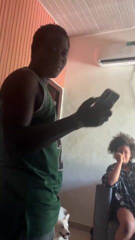 The video depicts a room with three individuals and a dog. A person in a green basketball jersey with "JORDAN 23" on the back stands in the foreground, looking at a smartphone. A girl with curly hair is sitting on a gray couch in the background. A white dog is visible near the legs of the person in the jersey. The room has an air conditioner mounted on the wall. The walls are painted in a light color, with a salmon-colored wooden panel on one side. The person with the phone walks around the room, briefly disappearing behind a dark shape and then reappearing in the frame.