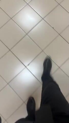 The video shows a first-person perspective of someone walking on a tiled floor. The person is wearing black pants and black shoes. The lighting in the scene is bright, casting reflections on the tiles. The camera is moving, indicating the person is in motion. The environment appears to be indoors, possibly a hallway or a public space with a grid-patterned tiled floor.