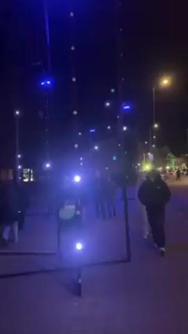 At night, people walk on a brick sidewalk alongside a street.  A black metal structure stands in the foreground, with vertical poles connected by horizontal bars near the ground.  Small white lights are attached to the structure, and blue lights shine from the top.  People walk between the poles of the structure. Streetlights illuminate the scene, and buildings can be seen in the background. A stroller is pushed along the sidewalk.  Cars drive on the street.