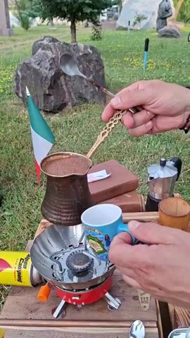 The video shows a close-up of hands pouring coffee from a copper cezve into a blue mug with a picture on it. The cezve is resting on a red portable camping stove. A decorative spoon with a gold handle is being used to assist in the pouring. To the left of the cezve, a small Italian flag is planted in the ground. In the background, there is grass, rocks, trees, and a statue. A yellow and red camping gas canister is visible on the left side of the frame.