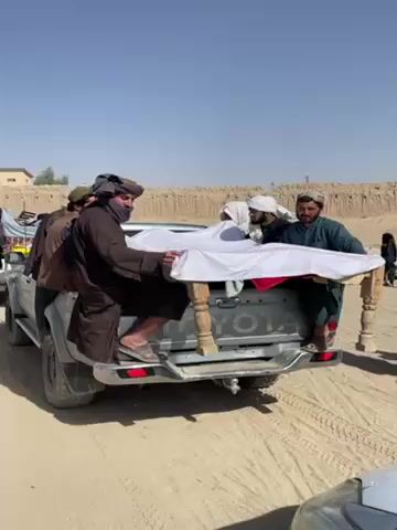 The video shows a group of men in a desert environment gathered around the back of a grey Toyota pickup truck. Four of the men are holding a wooden structure covered with white and red cloth in the truck bed. The men are wearing traditional clothing, and at least one man is wearing a face mask. Other people are present in the background. The sky is clear and blue. The ground is sandy and appears to be a dirt road. There is a low wall or embankment in the background.