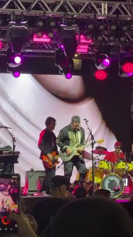 A live music performance is captured in a medium shot, focusing on a band playing on a stage.  The stage is illuminated by numerous purple and pink spotlights mounted on a metal truss structure overhead. A large screen behind the band displays a close-up image of a person's eye.  In the center foreground, a man wearing glasses and a dark jacket with ornate gold trim sings into a microphone while playing a silver electric guitar. To his left, another man plays a bass guitar and wears a red jacket. To the right, a drummer in a red hat and sunglasses plays a drum set. Several other band members or crew members are partially visible in the background. In the foreground, the heads and shoulders of audience members are visible, many holding up phones to record the performance. The overall atmosphere appears energetic and engaging.