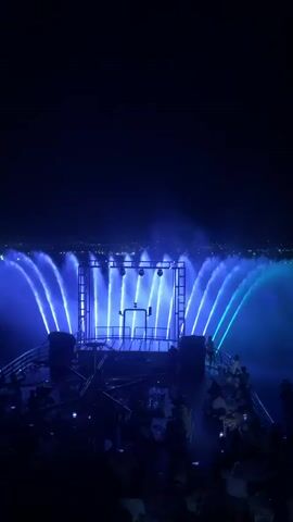 This is a night video of a synchronized water fountain and light show. The fountains are illuminated with changing colored lights, shifting from blue and green to yellow and red. In the foreground, there is a raised platform with what appears to be speakers and lighting equipment. A crowd of people is visible in the lower portion of the frame, many holding up their phones to record the spectacle. The background shows a dark sky with some distant city lights. The overall atmosphere is one of entertainment and visual spectacle.