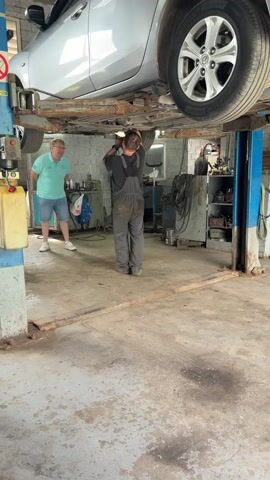 A man in overalls is working under a car that is raised on a lift. Another man in a light blue t-shirt and shorts is standing to the left, watching. The car is silver. The workshop floor is concrete and has some oil stains. There are tools and equipment visible in the background.