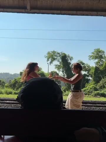 Two women playfully push against each other on a train platform, partially obscured by a large backpack in the foreground. They are standing beside train tracks on a sunny day, with lush green trees in the background. One woman wears a pink top, and the other is in a dark tank top and light pants. They are both smiling and interacting in a lighthearted manner. The platform is covered and shaded.