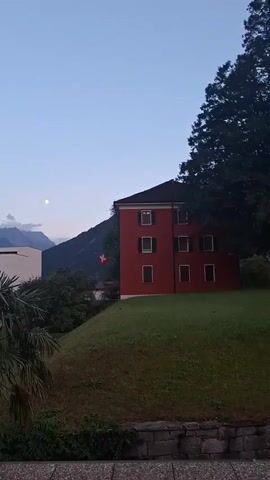 The video pans across a scene with buildings, trees, and mountains. In the foreground, there is a gray, textured surface, possibly a balcony or walkway. To the left, a palm tree is visible. Behind it, a modern white building with dark windows stands. A Swiss flag flies from a flagpole in front of the white building. Further to the right, a bright red building with multiple windows is situated on a grassy slope. In the background, large mountains dominate the landscape, and the sky is a pale blue, suggesting either dawn or dusk. The moon is also visible in the sky. The overall impression is a peaceful, possibly alpine setting at twilight.