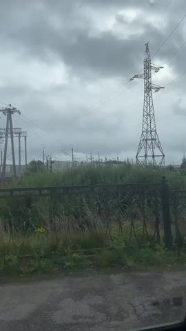 The video shows a view from inside a vehicle, looking out at a railway crossing. The scene is dominated by an overcast sky. In the foreground, a rough asphalt surface is visible, transitioning to a patch of grass with a black metal fence. Beyond the fence, there is overgrown vegetation and several utility poles with power lines. To the right, a large metal electrical tower stands prominently. Further in the distance, a small building with a red tiled roof and barred windows is visible, along with another small structure that appears to be a signal box. A blue barrel and some white bags are also present near the building. A railway crossing gate with red and white striped barriers is positioned across the road. The railway tracks themselves are visible stretching into the distance, with more overhead power lines and supporting structures. The overall impression is of an industrial or utilitarian area, possibly on the outskirts of a town or in a rural setting.