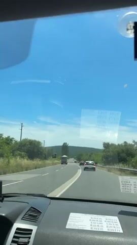 A first-person perspective video captures a view through a car windshield. The road ahead is visible with cars and a truck. The sky is bright blue with some clouds. The dashboard of the car is visible in the foreground, with a piece of paper with text and numbers on it. The landscape outside the car appears to be rural, with trees and fields. The video is likely taken while driving.