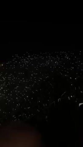 A high-angle view shows a concert venue at night, filled with a large crowd of people. The crowd is holding up lights, possibly cell phones, creating a sea of twinkling points. In the foreground is a bald head, belonging to a person presumably also attending the concert. As the video progresses, the camera pans slightly to reveal a stage with circular lighting elements and a large screen displaying a performer. There is a discernible figure on stage, though the details are not clear. A person in the foreground is holding up a phone to record the performance. The overall environment is dark, with the numerous lights held by the crowd and the stage lighting creating a vibrant contrast.