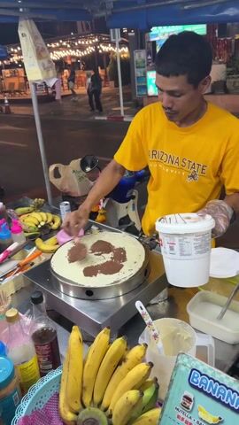 A street vendor in a yellow "Arizona State Sun Devils" t-shirt spreads Nutella on a crepe using a pink spatula. He stands behind a stainless steel cart with a round crepe griddle, bananas, and other ingredients and supplies. The background shows a street with pedestrians, vehicles, and street lights.