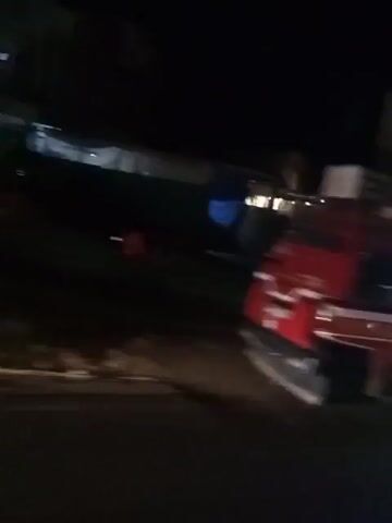 The video shows a night scene from a moving vehicle. It begins with a close-up of a red and white van with tinted windows. The camera then pans to show a street scene with several motorcycles parked and people walking around. There are shops and buildings visible in the background, some with lights on. The video ends with a shot of two white cars parked on the side of the road. The overall impression is of a busy street at night.
