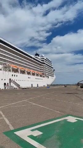 The video shows a large white cruise ship docked at a port. The ship has many decks with numerous windows and balconies. The hull of the ship has several round portholes. A gangway extends from the ship to the concrete pier. A marked helipad is on the concrete area next to the ship. In the background, a small group of people can be seen walking along the edge of the pier. The sky is blue with scattered white clouds.