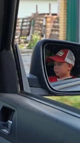 The video shows a young child wearing a red and white baseball cap and a red shirt reflected in a car's side mirror. The child is looking off to the side with a neutral expression. The interior of the car, including a door handle and a seatbelt, is visible in the foreground. The background outside the car consists of greenery and a fence, suggesting an outdoor setting on a sunny day. The car appears to be moving slowly or is stopped.