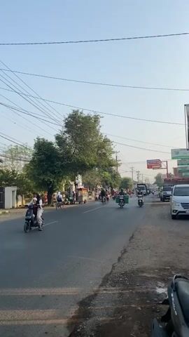 The video shows a street scene with various vehicles and people. On the left side of the road, there is a white building and a few trees. Several motorbikes are parked along the side of the road. A person on a white motorbike is visible, riding towards the camera. Further down the road, more people and motorbikes are present. The road itself is paved and appears to be a two-way street with traffic. On the right side of the road, there are shops and signs. A white van is parked on the right side. The sky is clear and blue. The video captures a busy street during the daytime.