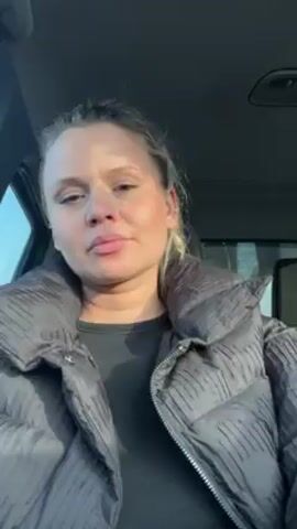 The video shows a woman inside a car. She has blonde hair pulled back, and is wearing a black top and a gray puffer jacket. The car's interior is visible, and there is sunlight coming through the window. The woman is looking at the camera and appears to be speaking.