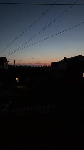 The video shows a twilight sky over a rural landscape with silhouetted houses and trees. A thin crescent moon is visible in the upper right portion of the frame. There are also two faint stars or planets visible in the upper left quadrant of the sky. Power lines are also visible crossing the sky. The overall atmosphere is dark and serene, suggesting the transition from day to night.