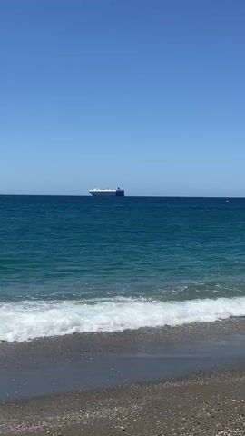 The video shows a clear, blue sky over a calm, turquoise ocean. A large, blue and white cargo ship is visible on the horizon. In the foreground, gentle waves are breaking on a sandy beach. The scene appears to be a sunny day.