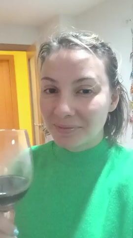 A woman in a green sweater is holding a wine glass filled with red wine. She lifts the glass to her lips and takes a sip. She is in a room with a white wall and a yellow door visible in the background.
