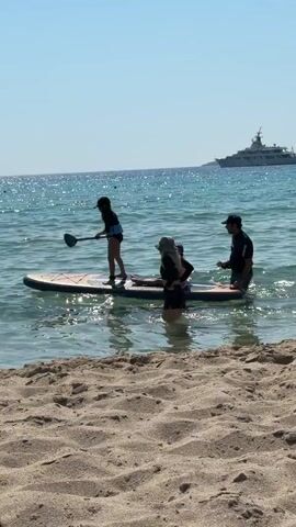 A paddleboard is in the shallow water near a sandy beach. There are four people on the paddleboard. One person is standing at the front and paddling. Two people are sitting in the middle, and one person is standing at the back. The water is clear and blue. A large white boat is visible in the background on the horizon. The sky is clear and blue.