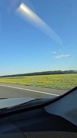 The video is captured from the perspective of someone inside a vehicle, looking out the windshield. The view is dominated by a clear blue sky with a few wispy clouds. A bright, elongated reflection of sunlight or a light source stretches across the upper part of the windshield. Below the sky is a flat, green landscape with a field of yellow flowers or crops stretching towards a treeline in the distance. A paved road is visible in the foreground, indicating the vehicle is in motion. The interior of the car is visible at the bottom of the frame, with a dark dashboard and speaker grille. The overall scene suggests a sunny day and a drive through a rural or open area.