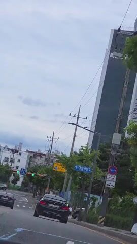 A low-angle, tilting shot captures a street scene from the perspective of a vehicle in motion. The camera pans upwards, revealing a cloudy sky, tall buildings, and utility poles with numerous wires. A dark-colored car is visible in the foreground, driving on the right side of the road, with other vehicles ahead and behind it. Streetlights and traffic signals are present, with some signals displaying green lights. Road signs are visible on poles, including a yellow sign with a blue silhouette of children walking and text in Korean, indicating a children's safety zone. Another blue sign with Korean text points to "Daejung Hospital." A tall, dark, slender building with Korean text on its upper portion looms in the background. Trees with green foliage line the right side of the road. A pedestrian can be seen walking further down the street.