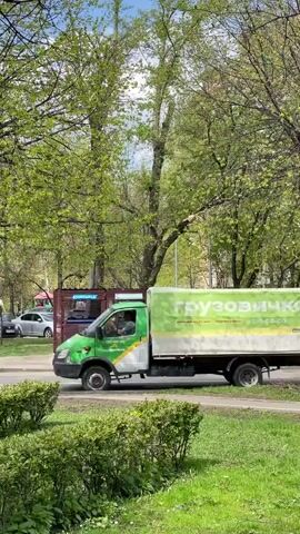 The video shows a static, medium shot of a street scene. On the left side of the frame, a green and white truck with the words "Грузовичкоф" in green letters drives away. The background features green trees with fresh spring leaves, and a brown corrugated metal structure that appears to be a kiosk. Several cars are parked nearby. The foreground includes a well-maintained green lawn with small, trimmed bushes. The day is bright and sunny.