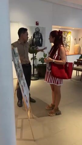 A man in a tan uniform is speaking to a woman with red hair, who is wearing a red and white striped dress and a red tote bag. They are standing in what appears to be a restaurant or cafe, with tables and chairs visible in the background. There is a display board in front of the man, and artwork on the wall behind him. A ceiling fan is visible above them.