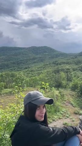 The video shows a landscape view from a high vantage point. A young woman with dark hair and a grey baseball cap initially stands in the foreground, looking towards the side, before the camera pans up to focus on the scenery. The primary subject is a lush, green mountain or hill, entirely covered with dense forest. The sky is overcast, with a mix of dark, storm-like clouds and patches of sunlight breaking through. The sun itself appears later in the shot, partially obscured by the clouds. The environment is natural, with sparse vegetation and a dirt path visible in the foreground, indicating a hiking area or trail. The overall mood is calm and serene, despite the somewhat ominous sky.