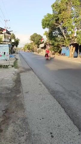 The video shows a paved road lined with trees and small buildings. A person on a pink scooter rides down the road from right to left. The Indonesian flag is visible flying from a pole. The sky is clear and blue. The overall impression is a sunny day in a village or small town.
