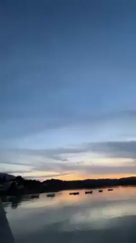 The video shows an airplane flying across the sky near the sea at dusk. The plane is heading toward the camera. The sky is blue and cloudy. The sea is calm and reflects the colors of the sky. There are some buildings on the shore and some boats on the water.