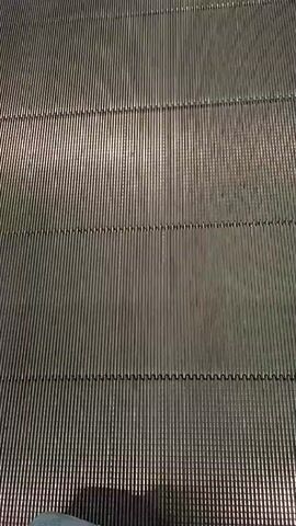 The video shows a first-person perspective of someone walking on a moving walkway in what appears to be an airport or large indoor public space. The walkway is made of metal slats that move horizontally. The environment is bright and modern, with a tiled ceiling and large glass windows on the left side. To the right, there are shops and people sitting or walking. The walkway is moving forward, and the camera is recording the path ahead. There are no other people on the walkway with the recording individual, but other people are visible in the background on the right side of the frame.