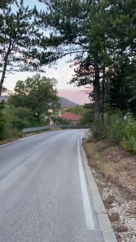 The video begins with a shot of a paved road winding through a wooded area. Tall trees line the right side of the road, and a guardrail is visible on the left. In the distance, a hazy pink and orange sky suggests either sunrise or sunset. The camera pans down to show a person’s legs and feet walking on the road. The person is wearing blue jeans and dark sneakers. The camera angle then shifts to a close-up of the road surface, showing cracked asphalt and a white dividing line. The person’s feet continue to walk forward, moving out of frame. The video ends with a blurry shot of the road ahead.