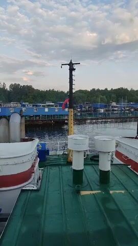 Here is a description of the video:

The camera is positioned on the green roof of a boat. In the foreground, there are two white and green cylindrical objects, along with lifeboats in white with red accents. In the background, across a body of water, there is a blue building with white windows and a red and white life preserver. A Russian flag is visible on a pole. Trees line the back of the blue building. The sky is partly cloudy.