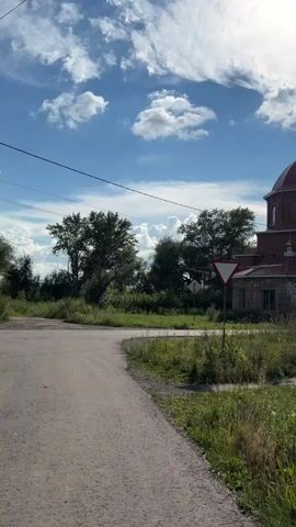 The video displays a rural scene with a brick church as the central focus. The church has a red roof and a golden dome topped with a cross. In front of the church, a light-colored car is parked. The surrounding environment is characterized by lush green grass and trees under a bright blue sky with scattered white clouds. A road curves through the foreground, and a yield sign is visible near the intersection. The sun is shining brightly, casting a warm light on the scene. The overall impression is one of a serene, sunny day in a tranquil, possibly historical, setting.