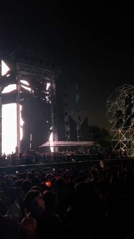 The video shows a large crowd of people at a concert at night. The crowd is facing a large stage with tall screens and scaffolding. The screens display various light patterns and images. The lighting on the stage is bright and dynamic. The sound of electronic music is audible. The environment appears to be outdoors and the mood is lively.