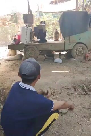 The video shows a man crouching in the foreground, facing away from the camera, looking at a machine on a trailer in the background. The man is wearing a dark blue shirt with white trim on the collar and a checkered baseball cap. He is also wearing yellow and black striped pants. He appears to be holding something in his hands, possibly a cigarette, and is making gestures with his hands. The trailer is loaded with a white bag, a black engine, and a green tarp covering the back. The trailer is resting on rocks. The environment appears to be outdoors, with dirt ground and some vegetation in the background. There is a rustic structure with a tarp overhead. The lighting suggests it is daytime.