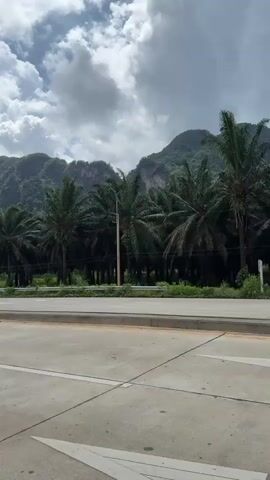 A POV video shows a car driving on a paved road, passing through an area with palm trees and karst mountains in the background. The sky is cloudy with some patches of blue sky visible. Traffic lights and streetlights line the road. There is a roundabout with a traffic island in the middle. A few cars and motorcycles are visible on the road. The video is shot during daylight hours.