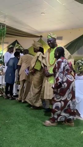 The video shows a room decorated with green and white drapery and faux greenery, suggesting an event or celebration. A group of people, predominantly dressed in traditional African attire in shades of beige and white with matching caps, are gathered. One woman in a long red patterned dress approaches a seated woman in a similar style of dress and appears to greet her with a bow. A man in a striped beige outfit stands nearby. The environment suggests a formal gathering or cultural event. The floor is covered in a green artificial turf. A few people are holding printed photos.