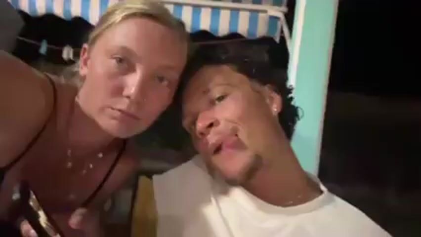 A blonde woman and a man with dark curly hair and a goatee make faces at a camera. They stick out their tongues and smile. The camera turns upside down and sideways. Behind them is a blue and white striped awning and a green pole. The man is wearing a white shirt. The background is dark.