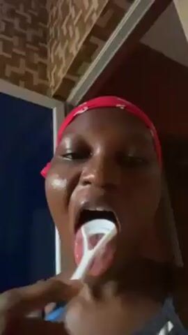A woman with dark skin and a red headscarf is shown in a close-up shot. She is facing the camera with her mouth wide open. She is using a white tongue scraper inside her mouth. Her teeth are visible. The background includes a blue door and a wall with a geometric pattern. The lighting is somewhat dim.