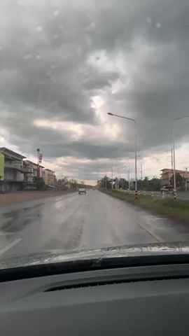 The video shows a view from inside a car driving on a wet road under a cloudy sky. The windshield has raindrops on it. The road appears to be a multi-lane highway with buildings and trees on the sides. There are a few other cars visible on the road ahead. The sky is filled with dark, heavy clouds, suggesting impending rain or a recent downpour. A windshield wiper is briefly visible sweeping across the windshield. The scene conveys a sense of travel during inclement weather.