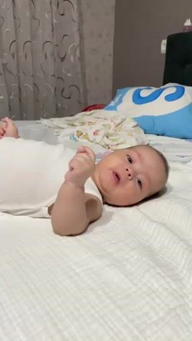 A baby is lying on a white, textured blanket in what appears to be a bedroom. The baby is wearing a white onesie and is looking at the camera with a curious expression, with their right hand near their mouth. In the background, there's a bed with a blue pillow that has a white "S" on it, and a white blanket with a floral pattern. There are also curtains in the background. The lighting suggests it might be daytime, but the room is not brightly lit.